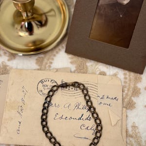 May include: An antique bronze chain bracelet rests on a vintage envelope with handwritten script and a postmark. A framed sepia-toned photograph and a gold candlestick with a lit candle are in the background.