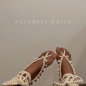 May include: Eggshell white crochet barefoot sandals adorned with seashells and small beads. The sandals are tied around the ankles and toes. The text "EGGSHELL WHITE" is displayed in the image.
