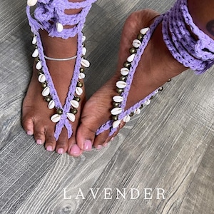 Puede incluir: Sandalias descalzas de ganchillo lavanda adornadas con conchas blancas y cuentas doradas. Las sandalias se envuelven alrededor de los tobillos y los dedos de los pies. La palabra "LAVENDER" est&aacute; impresa en blanco sobre la superficie de madera.