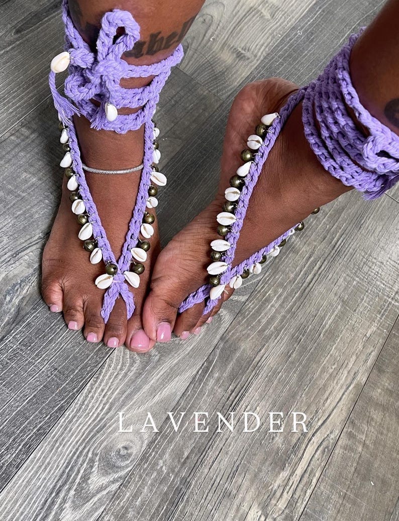 Puede incluir: Sandalias descalzas de ganchillo lavanda adornadas con conchas blancas y cuentas doradas. Las sandalias se envuelven alrededor de los tobillos y los dedos de los pies. La palabra "LAVENDER" est&aacute; impresa en blanco sobre la superficie de madera.