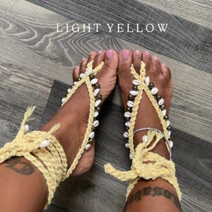 Puede incluir: Sandalias descalzas de ganchillo amarillo claro adornadas con conchas blancas y cuentas. Las sandalias se envuelven alrededor del tobillo y los dedos de los pies, creando un look playero y bohemio. El texto "LIGHT YELLOW" est&aacute; visible.