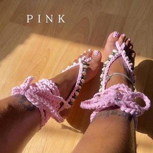 Puede incluir: Sandalias descalzas de ganchillo rosa adornadas con conchas marinas y cuentas. Las sandalias tienen un cierre de lazo alrededor del tobillo y se muestran sobre una superficie de madera. La palabra "PINK" se muestra en texto blanco.
