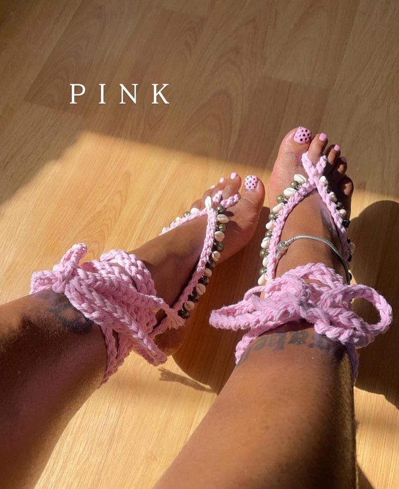 Puede incluir: Sandalias descalzas de ganchillo rosa adornadas con conchas marinas y cuentas. Las sandalias tienen un cierre de lazo alrededor del tobillo y se muestran sobre una superficie de madera. La palabra "PINK" se muestra en texto blanco.