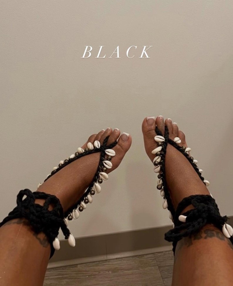 Puede incluir: Sandalias descalzas de ganchillo negro adornadas con conchas blancas y peque&ntilde;as cuentas. Las sandalias se envuelven alrededor del tobillo y los dedos de los pies, con un cierre de lazo. La palabra "BLACK" se muestra en texto blanco.