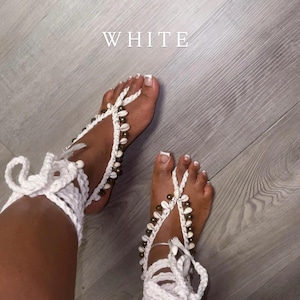 May include: White crochet barefoot sandals adorned with seashells. The sandals feature a woven design that wraps around the foot and ankle, with tie closures. The word "WHITE" is displayed in the image.