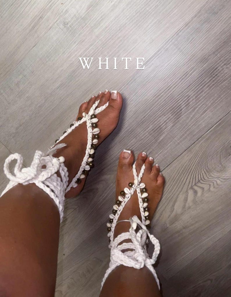 Puede incluir: Sandalias descalzas de ganchillo blanco adornadas con conchas marinas. Las sandalias presentan un dise&ntilde;o tejido que envuelve el pie y el tobillo, con cierres de lazo. La palabra "WHITE" se muestra en la imagen.