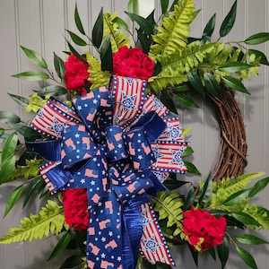 May include: A patriotic wreath featuring a grapevine base, adorned with red flowers, green foliage, and a large bow. The bow is made of blue and white ribbon with American flags and stars. A festive decoration for the home.