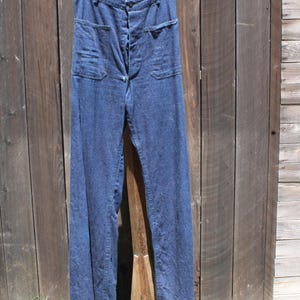May include: Dark blue denim pants with a button fly and two front pockets. The pants are hanging on a wooden stand against a weathered wooden background. The pants have a high waist and a wide leg.