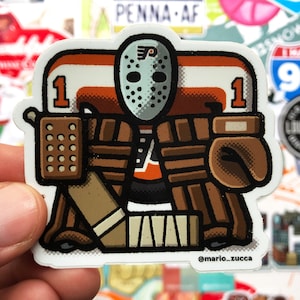 Può includere: Un adesivo con un portiere di hockey dei cartoni animati che indossa una divisa arancione, bianca e marrone con una maschera da hockey nera. Il portiere tiene un bastone da hockey e ha il numero "1" sulla sua maglia. L'adesivo ha il testo "@mario_zucca" in basso.