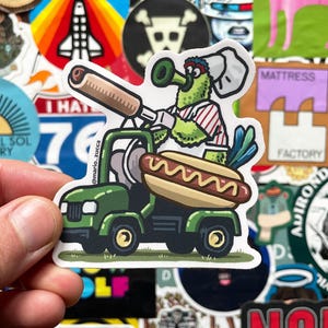 May include: A sticker featuring a cartoon character driving a green truck with a hot dog. The character is green with a chef's hat and a red and white striped shirt. The hot dog is in a bun. The sticker has the text "@mario_zucca".