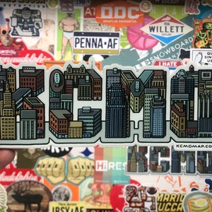Può includere: Un adesivo con un design di città stilizzato che compone la parola "KCMO" in uno schema di colori blu chiaro, grigio e marrone. L'adesivo ha uno sfondo bianco e un indirizzo del sito web in basso: "kcmopmap.com".