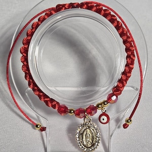 Peut inclure: Bracelet rouge tressé avec un pendentif doré de la Vierge Marie, entouré de cristaux transparents. Le bracelet comporte des perles rouges et un petit charme rouge en forme d'œil. Réglable avec des perles dorées.