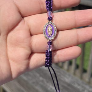 May include: A purple macrame bracelet featuring a central oval pendant with a gold-colored religious figure. The pendant is surrounded by clear crystals and a purple border. The bracelet has clear beads and a sliding knot closure.