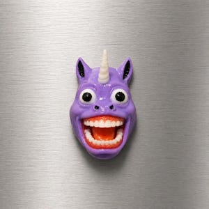 May include: A purple unicorn head with a wide, toothy grin, large black eyes, and a white horn. The ears have black detailing. The object is a novelty item, possibly a magnet or decoration.