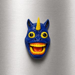 May include: A blue, cartoonish unicorn head with a yellow horn, large yellow eyes, and a wide, toothy grin. The mouth is open, revealing orange gums and a red tongue. The unicorn has black ears and is likely a magnet or decorative item.