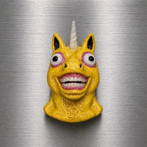 May include: A yellow unicorn head with a white horn, black ears, and large, wide eyes with pink outlines. The unicorn has a wide, toothy grin with pink gums and teeth. The object is likely a magnet or a decorative item.