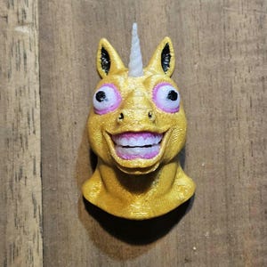 May include: A golden-yellow unicorn head with a white horn, large pink-rimmed eyes, and a wide, toothy grin. The unicorn has black details on its ears and a textured surface, creating a whimsical and slightly unsettling appearance.