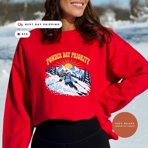 May include: A bright red crewneck sweatshirt featuring a graphic print of a skier on a snowy mountain with the text POWDER DAY PRIORITY in yellow arched lettering above a sunburst design, set against a backdrop of blue mountains and pine trees.