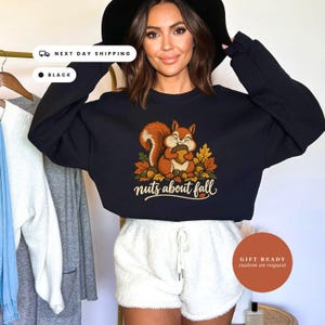May include: A black long-sleeve sweatshirt featuring a graphic of a brown squirrel holding an acorn surrounded by autumn leaves. The text "nuts about fall" is printed in a cream-colored script font below the squirrel illustration.