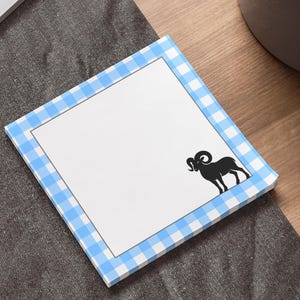 May include: Square notepad with a blue and white gingham border. A black silhouette of a ram is in the lower right corner. The notepad is on a wooden desk next to a laptop and a potted plant.