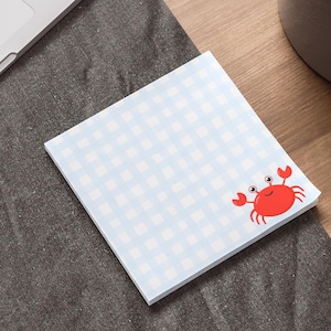 Puede incluir: Un bloc de notas cuadrado con un patrón de cuadros vichy azul claro y una ilustración de cangrejo rojo. El bloc de notas está sobre una superficie de madera, junto a un portátil y una planta en maceta.