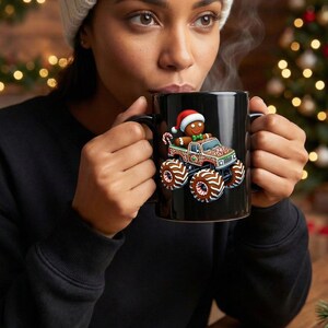 May include: Black ceramic mug with a festive gingerbread man in a Santa hat driving a monster truck. The truck and wheels are decorated with gingerbread-cookie-style designs. The mug has a black handle and interior.