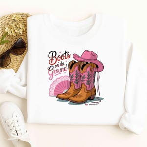 Peut inclure: Sweat-shirt blanc avec un motif de bottes de cowboy marron avec des détails floraux roses, un chapeau de cowboy rose et un éventail rose. Le texte "Boots on da Ground" est affiché en script noir et rose.