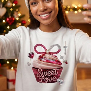 Peut inclure: Sweat-shirt blanc orné d'un motif de cupcake avec une crème au beurre rose et un bijou rouge. Le cupcake est orné d'un nœud rouge, d'un cœur et des mots "Sweet Love". Des ornements supplémentaires incluent une croix et un diamant.