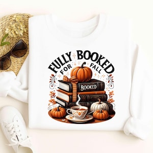 Puede incluir: Sudadera blanca con un diseño de temática otoñal. El diseño incluye una pila de libros, calabazas y una taza de té, con el texto "FULLY BOOKED FOR FALL". El diseño utiliza tonos naranja, marrón y negro.