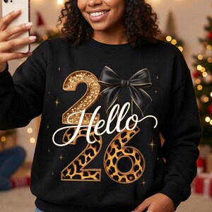 Puede incluir: Sudadera negra con el texto "Hello 26". El número "2" es dorado con purpurina, el número "6" tiene un diseño de estampado de leopardo y un lazo negro está encima del texto. La sudadera tiene cuello redondo.