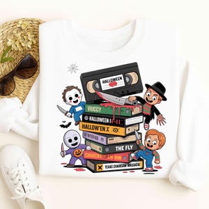 Puede incluir: Sudadera blanca con un diseño de personajes de películas de terror y títulos como "HALLOWEEN" y "The Fly" en libros apilados. Una cinta VHS está en la parte superior. El diseño incluye una telaraña y murciélagos.