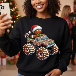 May include: Black sweatshirt with a gingerbread man wearing a Santa hat driving a monster truck. The truck has festive patterns and large, brown wheels. A candy cane is also visible.