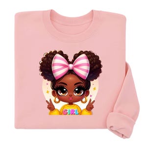 Peut inclure: Sweat-shirt rose clair avec une illustration de fille de dessin animé à la peau foncée, un nœud rose et blanc et le mot "GIRL" sur son haut jaune. La fille fait le signe de la paix. Boucles d'oreilles dorées également visibles.