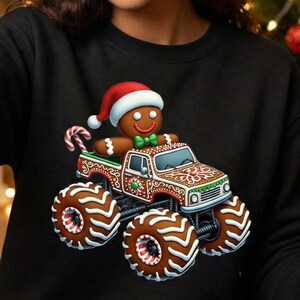 May include: Black sweatshirt with a gingerbread man in a Santa hat driving a gingerbread monster truck. The truck has large wheels and festive decorations. A candy cane is also visible.