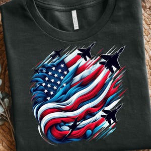 Könnte beinhalten: Dunkelgraues T-Shirt mit einem patriotischen Design. Die Grafik zeigt eine stilisierte amerikanische Flagge in Rot, Weiß und Blau, mit Kampfflugzeugen, die darüber fliegen. Das Design enthält Sterne und Streifen.