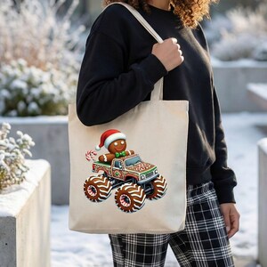 May include: A cream-coloured tote bag with a festive design. It features a gingerbread man in a Santa hat driving a decorated monster truck. The truck is brown, red, and green. The bag is held by a person, with a snowy background.