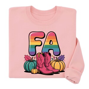 Peut inclure: Sweat-shirt rose clair avec les lettres "FA" aux couleurs de l'arc-en-ciel. Le motif comprend des bottes de cowboy roses, des citrouilles et des feuilles d'automne. Le sweat-shirt est de style col rond.