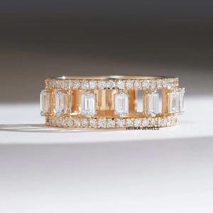 May include: A gold-toned ring featuring a series of rectangular, clear gemstones set between rows of smaller, round, clear stones. The ring has a band with an open design, and the text "HITIKA JEWELS" is visible.