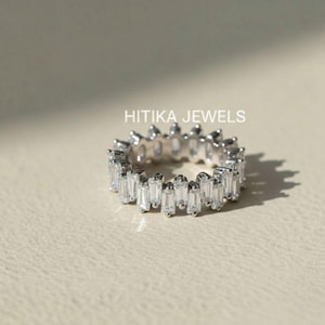May include: A silver ring with a continuous band of rectangular, clear gemstones. The ring is set on a neutral-toned surface, with the text "HITIKA JEWELS" visible in the background.