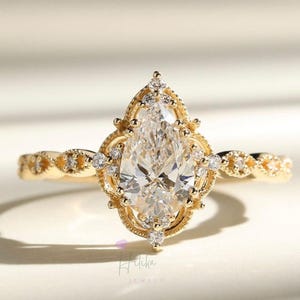 May include: A gold ring featuring a pear-shaped diamond center stone. The ring has a detailed filigree design around the diamond and smaller accent stones. The band has a delicate, twisted pattern. The ring is a piece of jewelry.