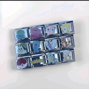 May include: A collection of silver-toned charm links featuring colorful cartoon character designs. The charms include various creatures and a Pokeball, stacked on a white surface. The charms are rectangular and have a glossy finish.