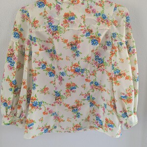 May include: A cream-colored long-sleeve blouse featuring a vibrant floral pattern in shades of blue, orange, and pink. The garment has a high neckline with two symmetrical cut-out details near the collar and full sleeves with buttoned cuffs.