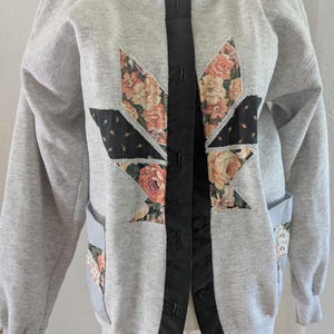 May include: A gray sweatshirt jacket with floral patchwork details. The jacket features a black satin trim down the front with button closures and pockets. The floral patches include roses and other flowers in shades of pink, peach, and cream.