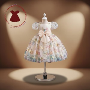 Könnte beinhalten: Ein weißes Kleid mit floralem Muster, einer großen Schleife und Puffärmeln. Das Kleid hat einen weiten Rock mit farbenfrohem Blumenmuster und goldenen Akzenten. Das Kleid wird auf einer Schaufensterpuppe präsentiert.