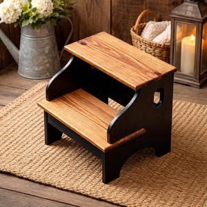 May include: A two-step wooden stool with a natural wood top and step, and black supports. The stool has a handle cut out on the sides. The stool is placed on a woven jute rug, with a rustic background.