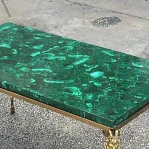 以下が含まれることがあります： マラカイトの天板と金色の金属フレームを備えた長方形のコーヒーテーブル。天板は、濃い緑色の渦巻き模様が特徴的な鮮やかな緑色です。脚には装飾的なディテールがあり、デザインにエレガンスを加えています。テーブルの長さは約120cmです。