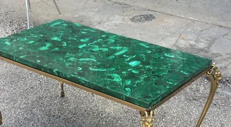 以下が含まれることがあります： マラカイトの天板と金色の金属フレームを備えた長方形のコーヒーテーブル。天板は、濃い緑色の渦巻き模様が特徴的な鮮やかな緑色です。脚には装飾的なディテールがあり、デザインにエレガンスを加えています。テーブルの長さは約120cmです。
