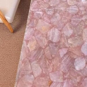May include: A close-up of a table surface made of pink rose quartz. The surface is composed of irregular, interlocking pieces of the stone, creating a mosaic-like pattern. The color palette is soft, with variations of pink and white.