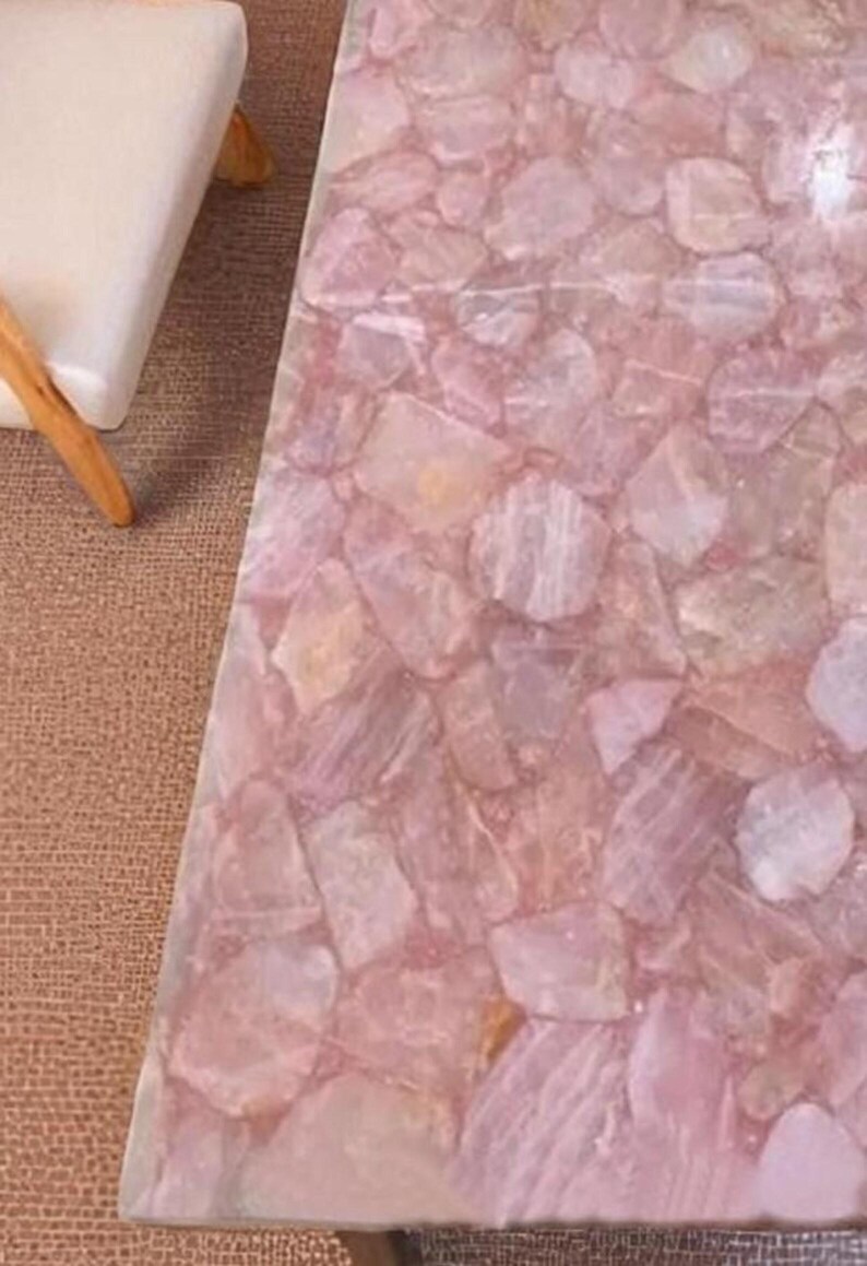 May include: A close-up of a table surface made of pink rose quartz. The surface is composed of irregular, interlocking pieces of the stone, creating a mosaic-like pattern. The color palette is soft, with variations of pink and white.
