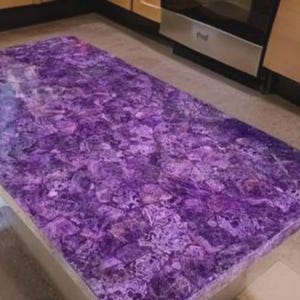 Puede incluir: Una encimera rectangular de amatista con una superficie pulida. La encimera es de color p&uacute;rpura oscuro con vetas p&uacute;rpuras y blancas m&aacute;s claras. La encimera est&aacute; colocada en una cocina con gabinetes de color claro y electrodom&eacute;sticos de acero inoxidable.